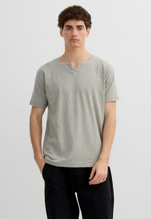 Joven con cabello corto y castaño que lleva una camiseta gris claro de manga corta con cuello henley y pantalones negros, sobre un fondo liso.