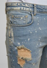 Svijetloplave traperice s izblijedjelim završetkom, uzorkom prskanja i istrganim koljenom koje ima razderotine i izlizane rubove.