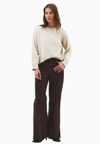 Maglione color crema con ampi polsini, caratterizzato da dettagli con bottoni sulle spalle, abbinato a pantaloni svasati marroni a vita alta.