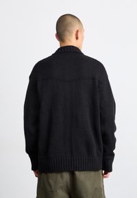 Cardigan nero lavorato a maglia con orlo e polsini a coste, caratterizzato da un colletto a scialle e un motivo a punto testurizzato, visto da dietro.