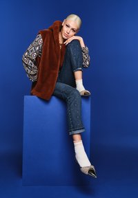 Bruine nepbont gilet over een gestreept shirt, denim jeans met versieringen, en sprankelende puntige schoenen gedragen met witte sokken.