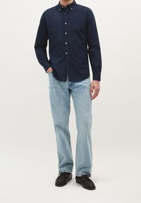 Marineblaues Hemd mit Knöpfen, Kragen und einer einzelnen Brusttasche, kombiniert mit hellblauen Jeans und dunklen Schuhen. Die Struktur wirkt glatt und lässig.