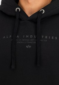 Sudadera negra de algodón con capucha ajustable. Presenta un logo y texto impresos en el pecho, y ojales plateados para los cordones.