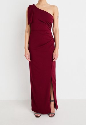 Kobieta ubrana w bordową, asymetryczną, długą suknię z rozcięciem z boku oraz czarne buty na wysokim obcasie z odkrytymi palcami, stojąca na tle jednolitej białej ściany.