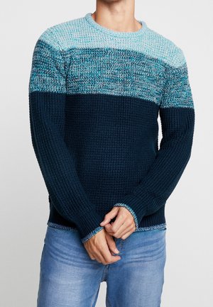 Mand iført en tyk strikket sweater med lyseblå, turkise og marineblå farveblokke, kombineret med lyseblå jeans, hænderne foldet foran.