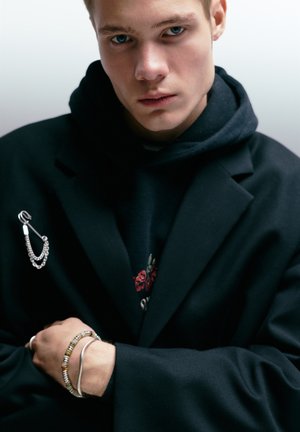 Zwarte dubbelreeks jas over een zwarte hoodie, met een zilveren kettingbroche en gelaagde armbanden met metalen en kleurrijke kralen.