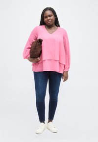 Γυναίκα με μακριά πλεξούδες, φοράει ροζ μπλούζα με στρώσεις, σκούρο μπλε τζιν και λευκά αθλητικά παπούτσια, κρατώντας ένα διπλωμένο καφέ σακάκι.