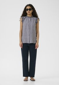 Hele hallist ilma varrukateta pluus, millel on Volangõlad ja nööbid esiosas; kombineeritud tumesiniste laia jala pükstega ja mustade sussidega.