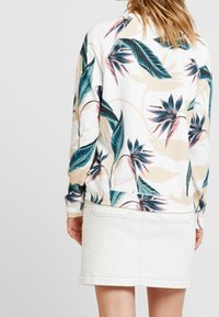 Sudadera con estampado floral en tonos verde azulado, rosa y beige sobre tela blanca; presenta puños acanalados. Combinada con una falda de mezclilla blanca.
