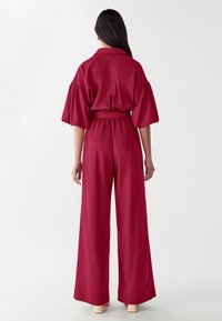 Raudonas kombinezonas su apykakle ir pūstomis rankovėmis. Medžiaga lygi, su diržu juosmenyje ir plačiomis kelnėmis, tinka kasdieniam dėvėjimui.