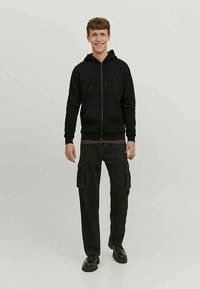 Giovane uomo sorridente, che indossa una felpa nera con zip, pantaloni cargo neri e scarpe nere, in piedi contro uno sfondo semplice.