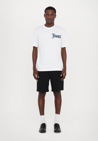 Biela bavlnená tričko s čiernym nápisom "Just", skombinované s čiernymi kraťasmi. Model má na sebe biele ponožky a čierne topánky. Jednoduchý, casual outfit.