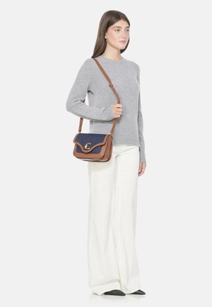Borsa a tracolla in pelle marrone con pattina blu, hardware dorato e design curvo. Modella in maglione grigio e pantaloni bianchi si trova accanto.