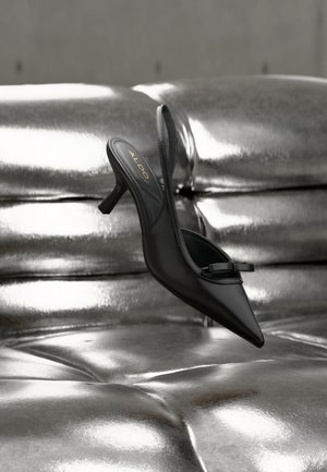 Chaussure à talon noire à bout pointu et bride arrière, avec un petit nœud, reposant sur un revêtement en cuir argenté brillant matelassé.