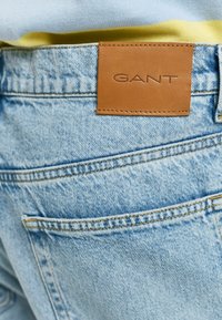 Denim jeans with a light blue wash, featuring a brown leather label embossed with "GANT" on the back pocket, showcasing sturdy stitching.