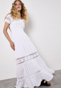 Witte maxi-jurk met een aansluitend lijfje, pofmouwen en een smocktail. Bevat bloemenkant accenten en een gelaagde rok met een gerimpelde zoom.