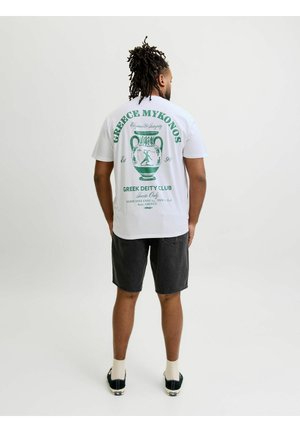 Muškarac s dredovima nosi bijelu grafičku majicu sa zelenim grčkim vazu dizajnom i crne kratke hlače, stoji na jednobojnoj pozadini.