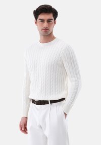 Weißer gestrickter Pullover mit einem Zopfmuster, Rundhalsausschnitt, Rippbündchen und einer taillierten Form, kombiniert mit hellen Hosen und einem braunen Gürtel.