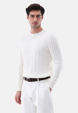 Maglione bianco lavorato a maglia con motivo a treccia, scollo rotondo, polsini a coste e forma aderente, abbinato a pantaloni chiari e una cintura marrone.