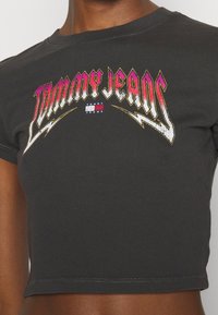Černé zkrácené tričko s ribbovanou texturou. Obsahuje barevnou grafiku "Tommy Jeans" s blesky v růžové, zlaté a bílé barvě.