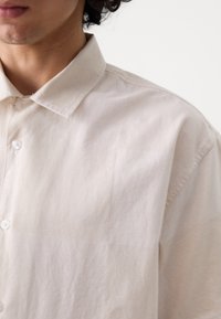 Buste d'une personne portant une chemise beige clair à boutons avec un col, présenté sur un fond blanc uni.