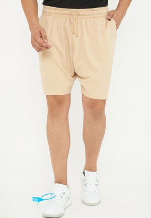 Uomo che indossa pantaloncini beige con coulisse e tasche laterali e sneakers bianche con etichetta blu sulla scarpa sinistra, in piedi davanti a uno sfondo bianco.