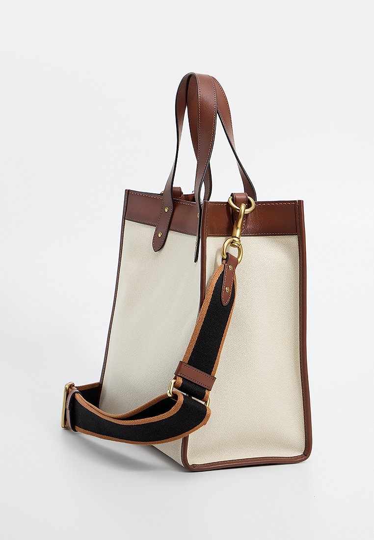 Bolso de lona con detalles y asas de cuero marrón. Presenta una correa negra desmontable y herrajes de oro. Forma rectangular, abertura superior.