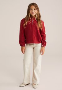 Maglietta rossa a maniche lunghe con tessuto testurizzato, colletto abbottonato e dettagli di cucitura bianca; abbinata a pantaloni larghi beige chiaro e scarpe color crema.
