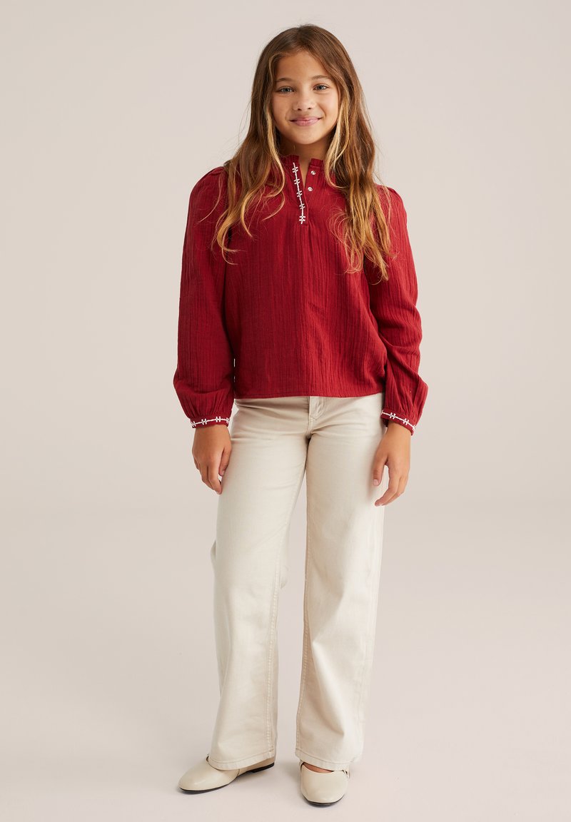 Maglietta rossa a maniche lunghe con tessuto testurizzato, colletto abbottonato e dettagli di cucitura bianca; abbinata a pantaloni larghi beige chiaro e scarpe color crema.