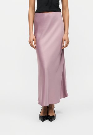 Frau trägt einen hoch taillierten, seidigen Maxi-Rock in helllila, kombiniert mit schwarzen spitzen Schuhen und einem schwarzen, kurz geschnittenen Oberteil.