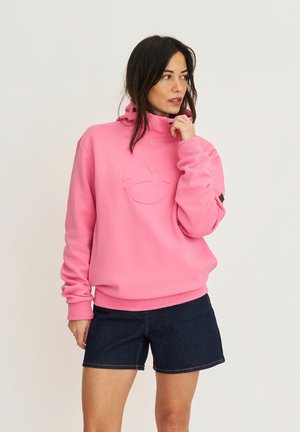 Femme aux longs cheveux foncés portant un sweat à capuche rose et un short en jean foncé, regardant pensivement sur le côté sur un fond uni.