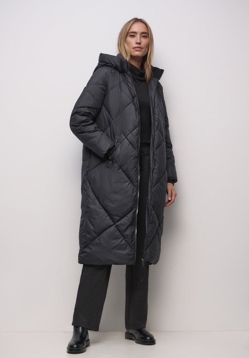 Nainen seisoo pukeutuneena pitkään mustaan tikattuun takkiin, jossa on huppu, mustiin housuihin, mustiin saappaisiin ja mustaan toppiin yksiväristä taustaa vasten.