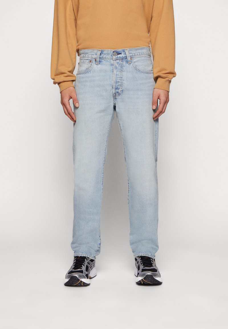 Hellblaue Denim-Jeans mit geradem Schnitt, Fünf-Taschen-Design und sichtbarem Nähen. Kombiniert mit einem beigen Langarmshirt und schwarzen Sneakers.