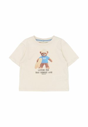 Camiseta de manga corta de color beige claro, con un gráfico de un oso con una camiseta azul sosteniendo una tabla de surf, con el texto: "Esperando esa ola perfecta".