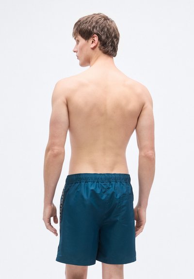 Ανδρικά μαγιό σε σκούρο τιρκουάζ, κατασκευασμένα από γυαλιστερό ύφασμα. Διαθέτουν ελαστική μέση και πλαϊνή τσέπη. Απλός, λειτουργικός σχεδιασμός.