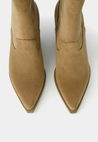 Beżowe botki zamszowe za kostkę z ostro zakończonym noskiem, z minimalnymi przeszyciami i wzmocnionym obcasem. Gładka faktura i elegancki design.