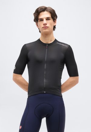 Νέος άνδρας που φοράει μαύρο ποδηλατικό μπλουζάκι με κοντά μανίκια και μπλε σκούρο επενδυμένο ποδηλατικό σορτς, στέκεται με τα χέρια πίσω από την πλάτη.