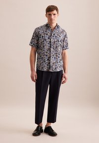 Camicia a maniche corte con bottoni, caratterizzata da un motivo floreale multicolore su uno sfondo scuro, abbinata a pantaloni blu scuro e scarpe nere.