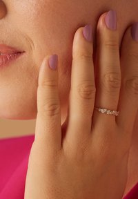 Bague en or avec plusieurs petites pierres claires, présentée sur une main à la peau claire et aux ongles manucurés couleur lavande, touchant la joue.