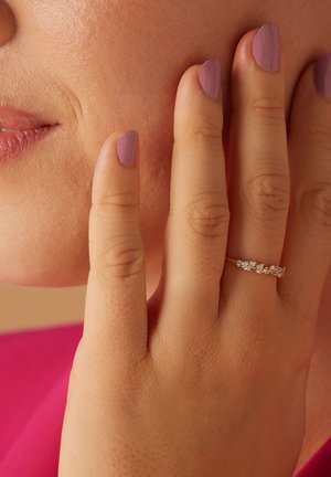 Bague en or avec plusieurs petites pierres claires, présentée sur une main à la peau claire et aux ongles manucurés couleur lavande, touchant la joue.
