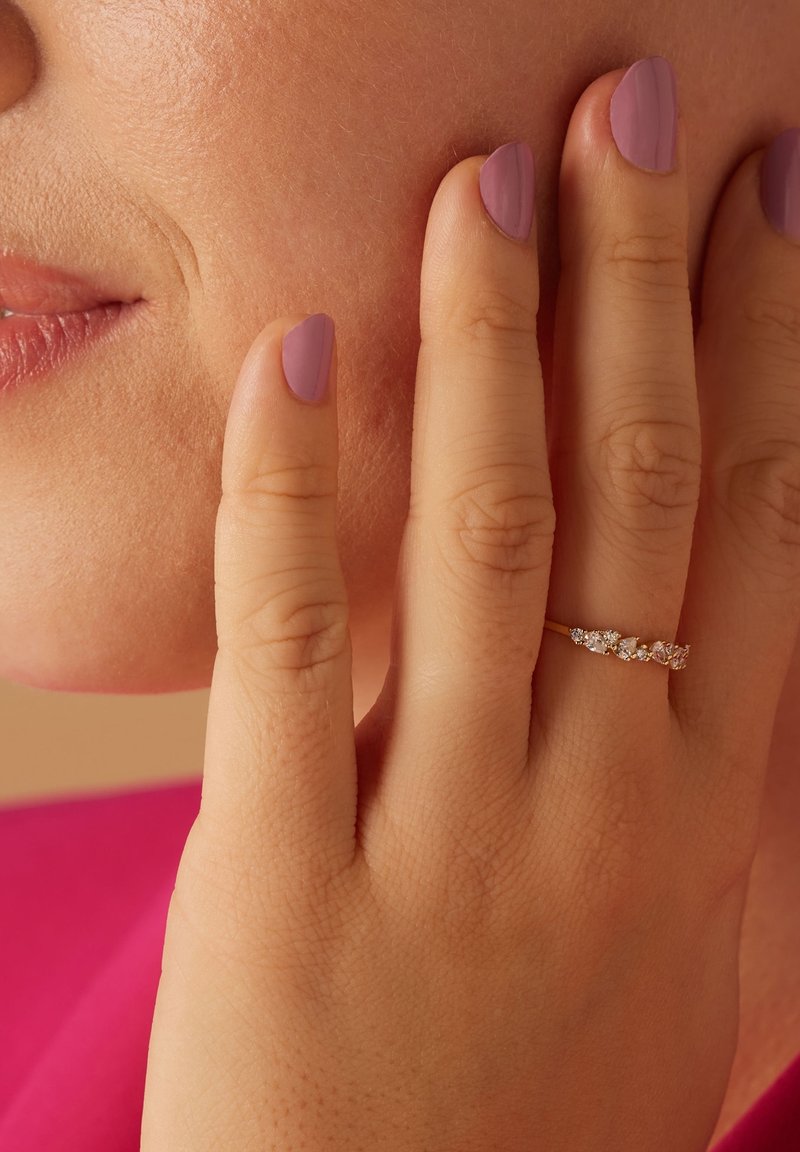 Gouden ring met meerdere kleine heldere edelstenen, weergegeven op een hand met lichte huid en gemanicuurde lavendel nagels, die de wang aanraakt.