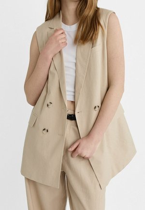 Femme portant un blazer beige sans manches à double boutonnage, un pantalon assorti et un débardeur blanc court, tenant le revers du blazer d'une main.
