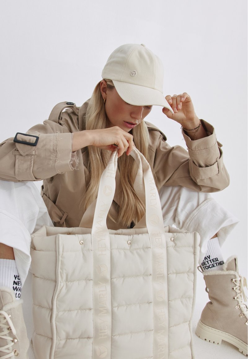 Grote beige gewatteerde tote bag met crèmekleurige banden, met subtiele branding. Het model draagt een bijpassende beige pet en lichte kleding.