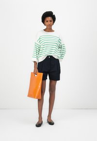 Übergroßer weißer Pullover mit grünen Streifen, marineblaue Tailored-Shorts und eine orange Tote-Tasche, kombiniert mit schwarzen flachen Schuhen.