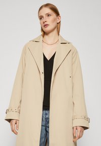 Beige trenchcoat med brede revers, knappdetaljer og justerbare mansjetter. Båret over en svart topp og parret med blå jeans.