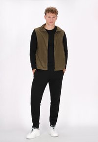 Olivgrüner Zip-Fleece-Westen über einem schwarzen Langarmshirt, kombiniert mit schwarzen Jogginghosen und weißen Sneakern. Glatte Textur, lässige Passform.