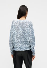 Γυναίκα με μακριά σκουρόχρωμα μαλλιά που φοράει πουλόβερ με λεοπάρ σχέδιο σε μπλε και άσπρο και μαύρο παντελόνι, εμφανιζόμενη από πίσω μπροστά σε ένα μονόχρωμο φόντο.