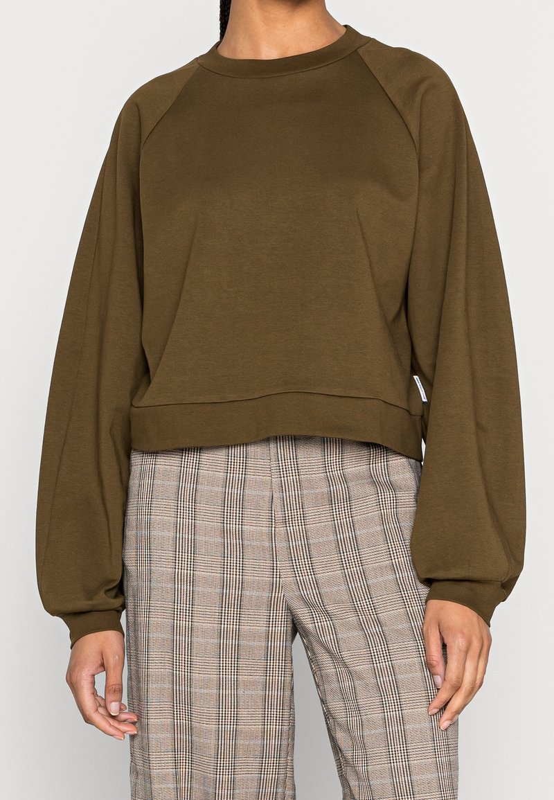 Oliwkowa krótsza bluza z raglanowymi rękawami i gładką fakturą. Połączona z wysokim stanem, beżowo-czarnymi, kratkowanymi spodniami.