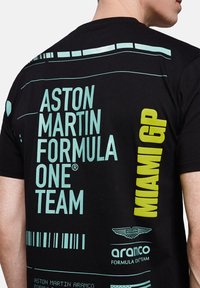 Czarny t-shirt z turkusowym i limonkowym tekstem. Wzory obejmują "Zespół Formuły 1 Aston Martin", "Miami GP" oraz logotypy. Materiał bawełniany.