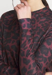 Långärmad topp i mörk burgundy med svart leopardmönster. Ribbad textur och figurnära design med hög krage.
